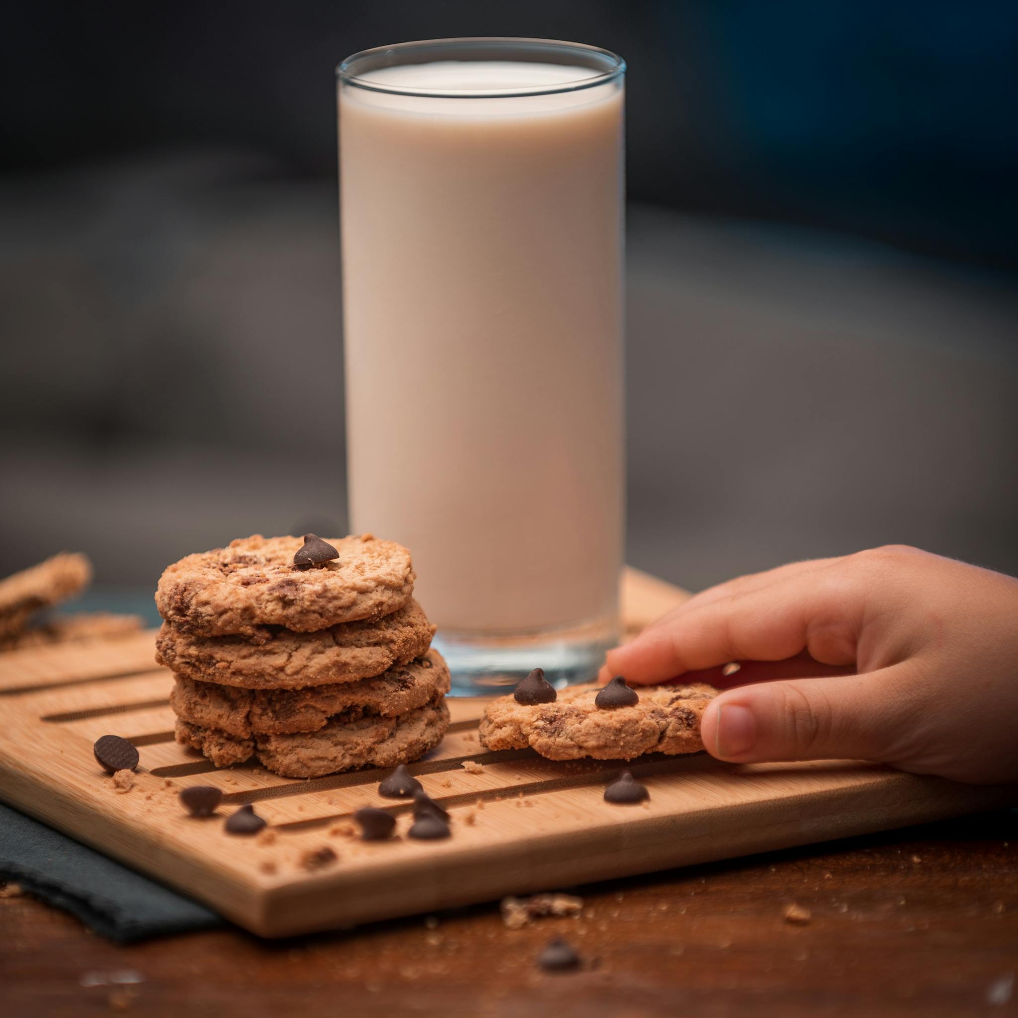 A close-up of chocolate chip cookies and a glass of milk on a wooden tray, with a child's hand reaching.